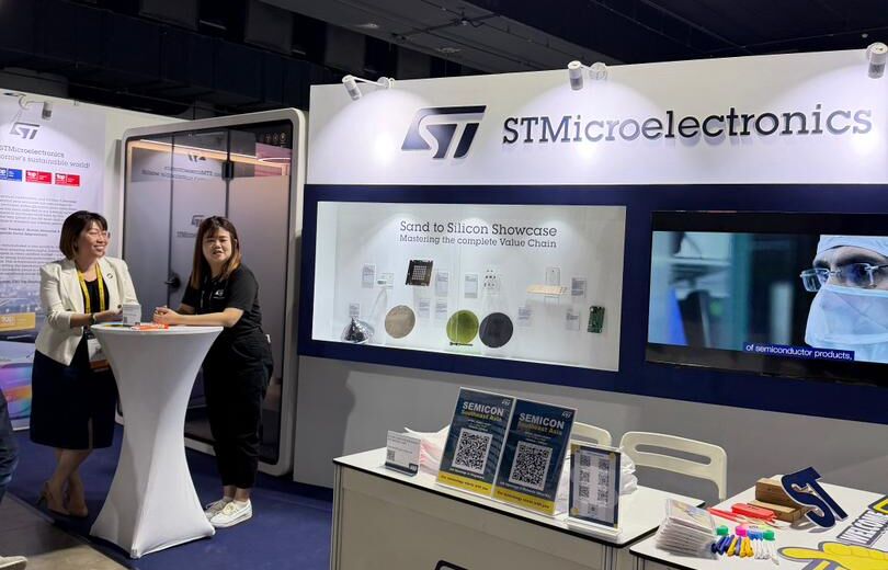 Two women chatting beside a round high table next to an acoustic booth at the STMicroelectronics exhibition stand during SEMICON 2026