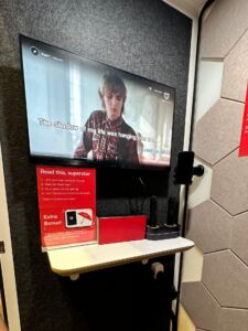 Wall-mounted TV screen inside an acoustic booth displaying karaoke lyrics, with a microphone stand and red instructional signage on a shelf below