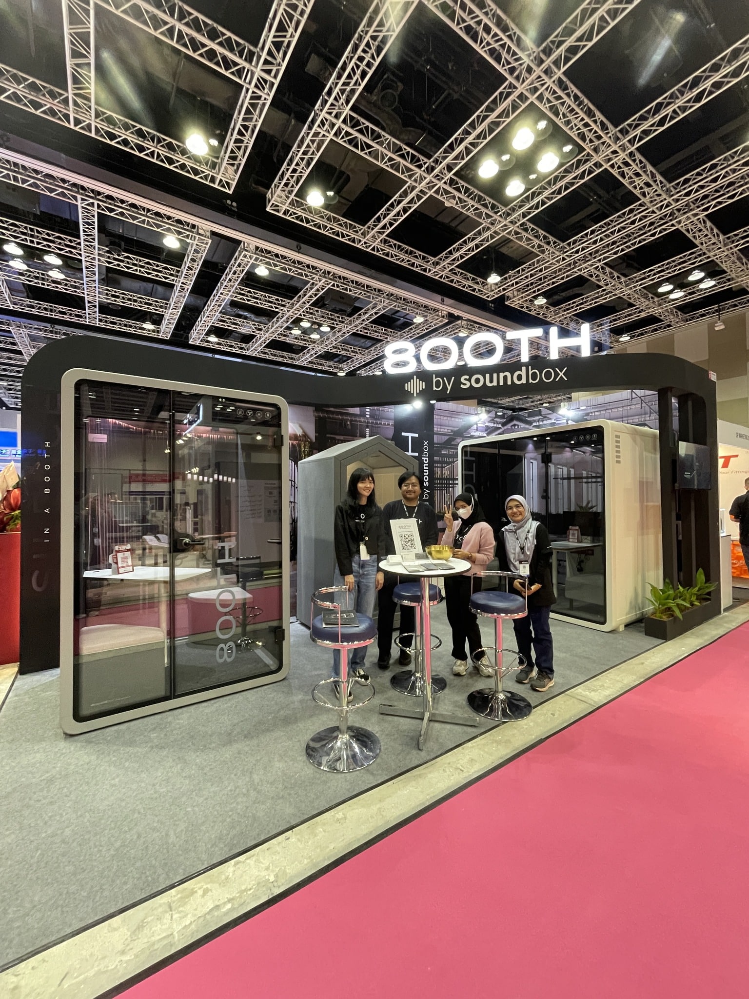 800TH by Soundbox exhibition booth at Archidex, with a glass-panelled acoustic pod on the left and two team members posing with two visitors at a high table between two booths