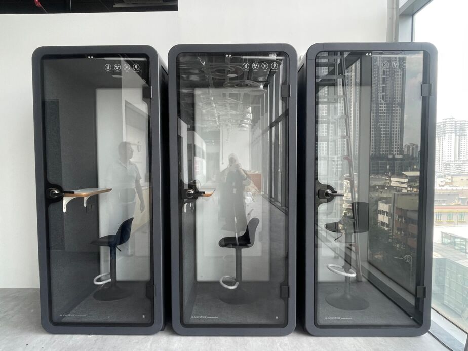 Three grey acoustic booths with bar stools in a row beside a window with a city view