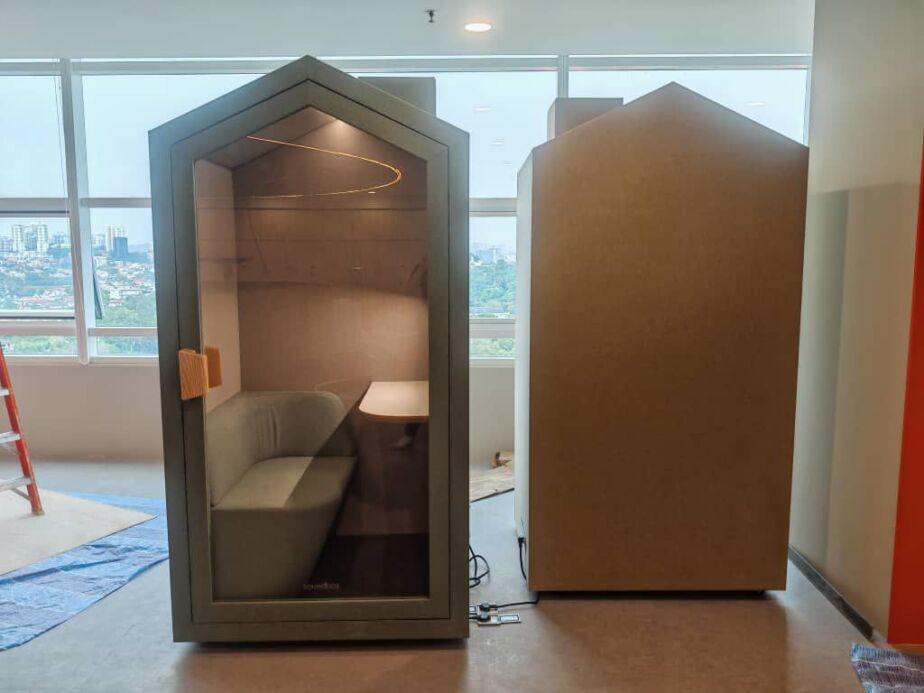 Pitched-roof acoustic booth with sofa and desk beside a plain fabric-panel booth, in front of floor-to-ceiling windows
