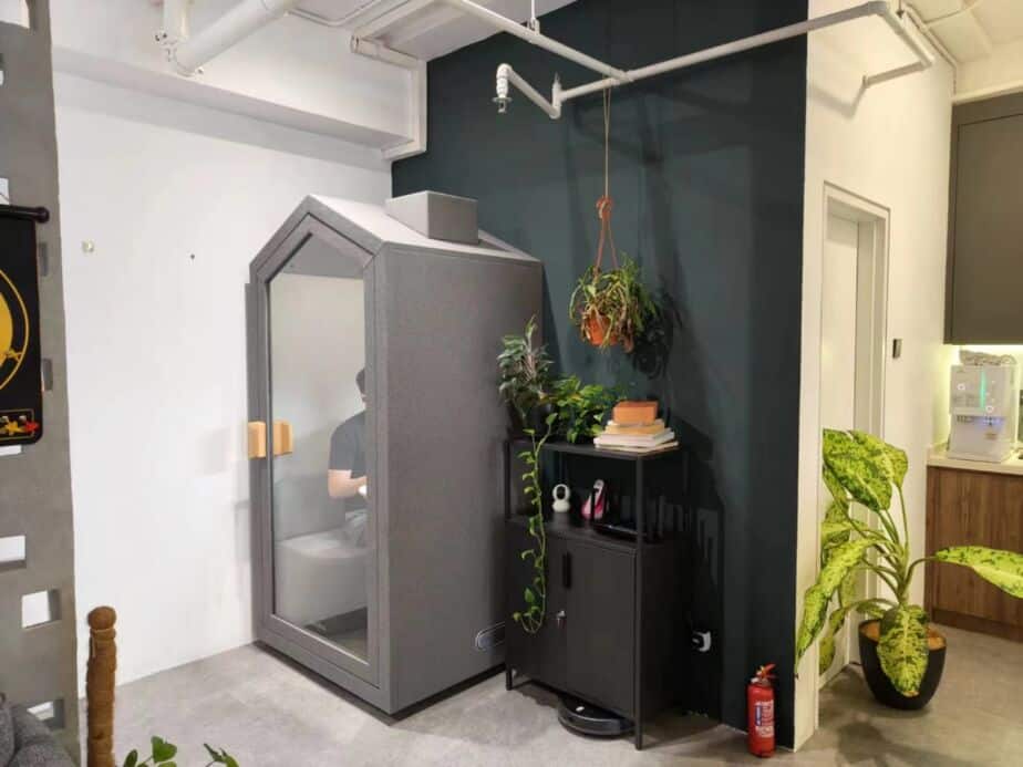 Grey pitched-roof acoustic booth in an office corner with a person seated inside, next to potted plants